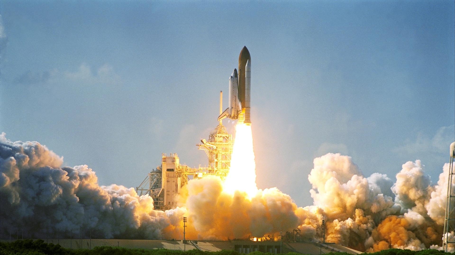 an orange rocket launches above a plume of flame and smoke, carrying a white aircraft on its back