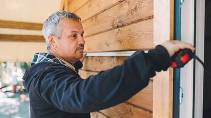 An older man uses a tape measure next to the door on the outside of a cabin.