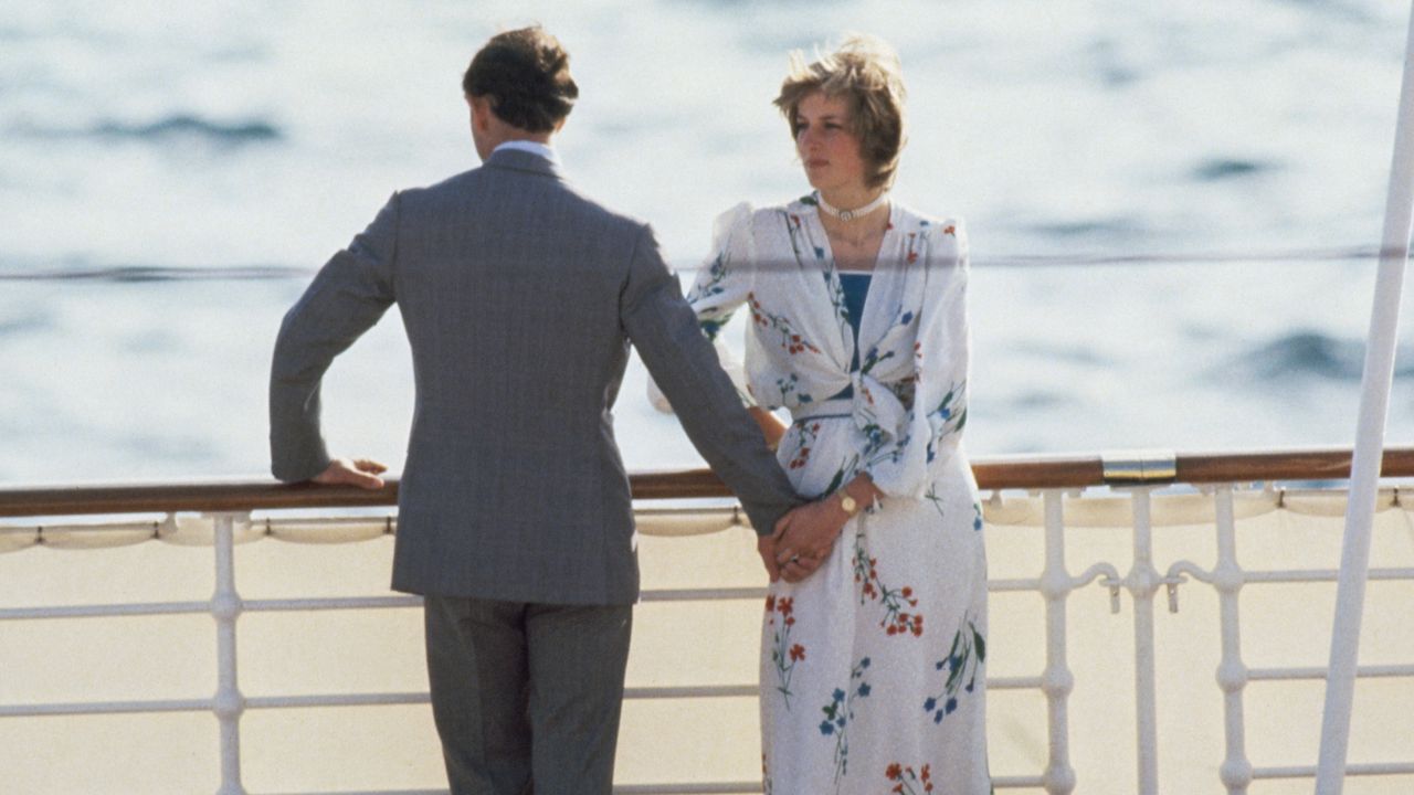 Princess Diana wearing a white floral dress standing on a ship with her back to the water while Prince Charles, wearing a gray suit, is turned the other way and holding her hand