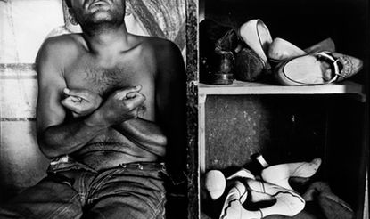 Black and white image of a topless seated male leaning his head back against a stone wall, arms crossed over and hands clenched, wearing open waisted jeans, to the right side of him an old shelving unit containing ladies heeled worn shoes scattered on the shelves