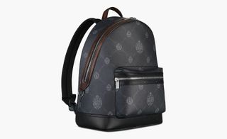 View of a black and brown Berluti backpack featuring a repeating pattern made up of the brand logo and a front compartment with a zip. The backpack is pictured against a light coloured background