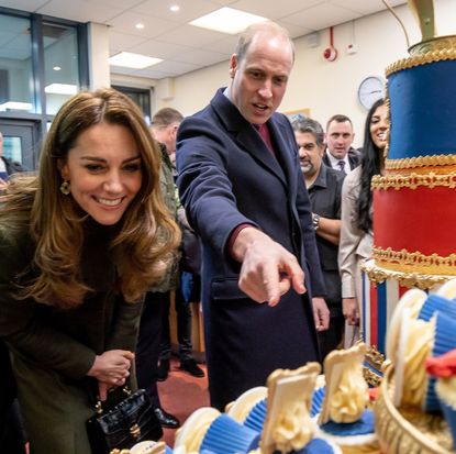 Prince William pointing at a blue and white cupcake and Princess Kate leaning forward to look at it and smiling