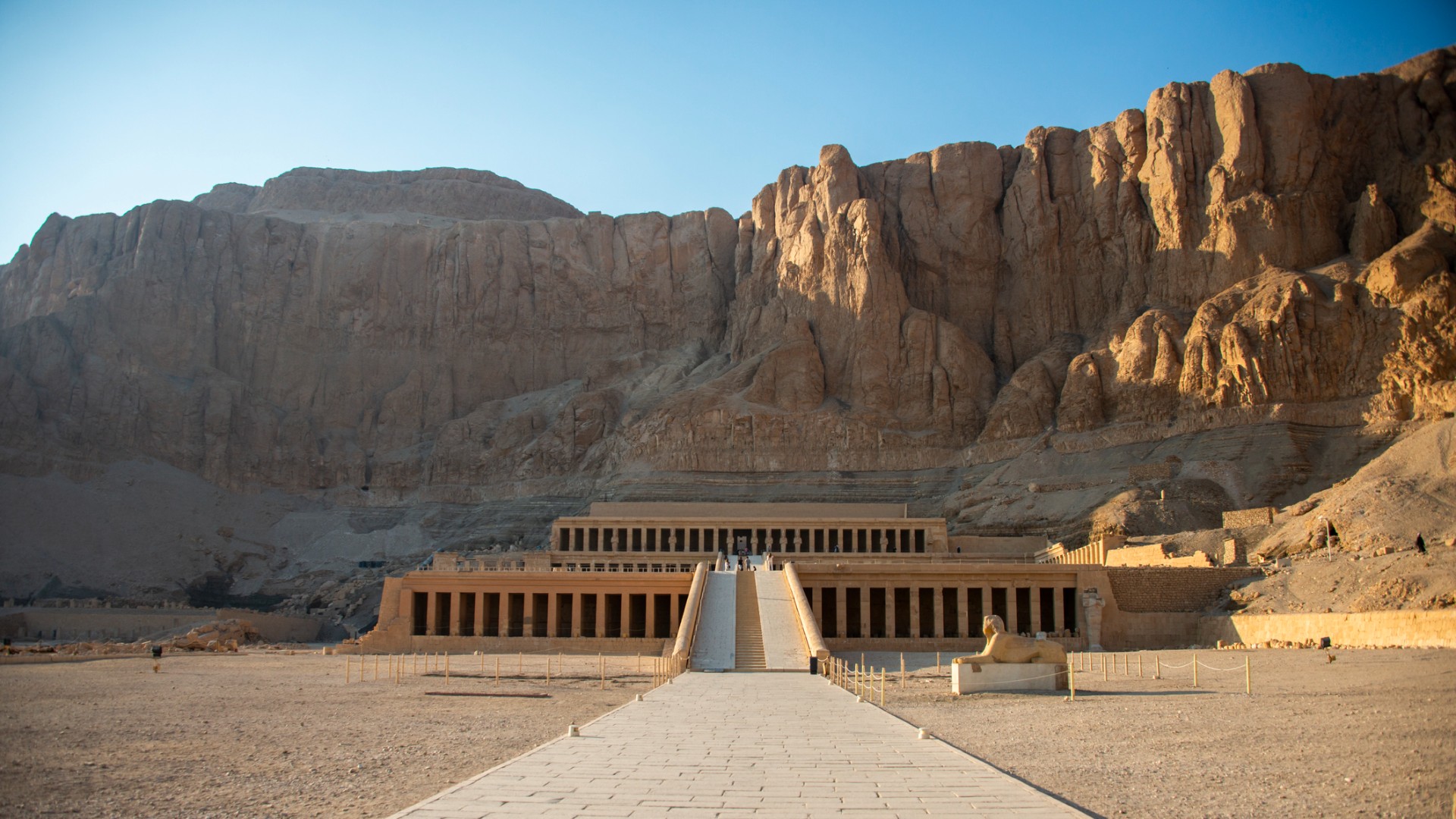 O antigo templo egípcio de Hatshepsut fica no final de uma longa passarela de pedra sob imponentes penhascos do deserto, evocando uma grandeza tranquila e iluminada pelo sol.