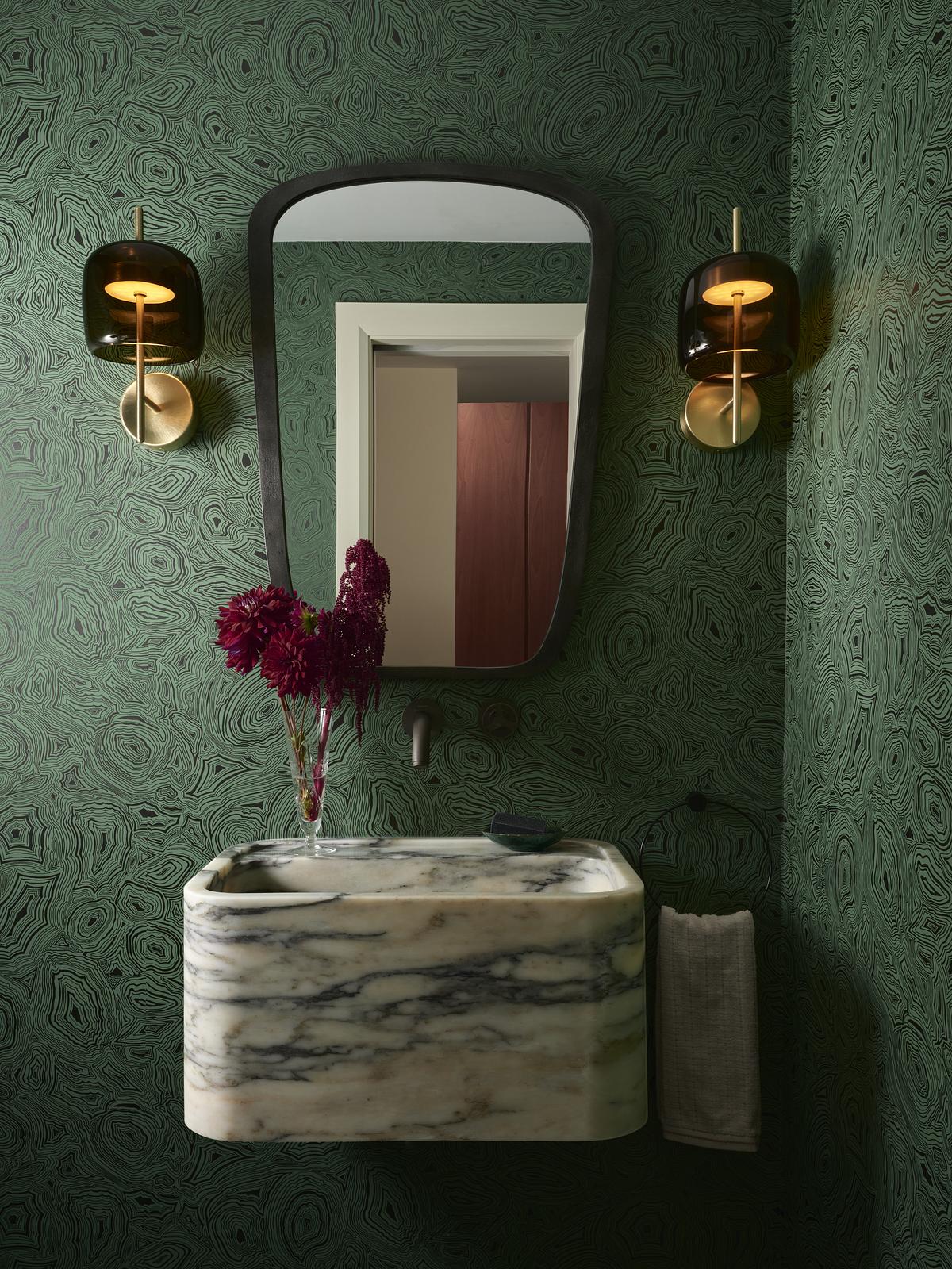 A green powder room with patterned wallpaper, a floating marble sink with a vase of flowers styled on it, a round mirror on the wall with wall sconce on either side