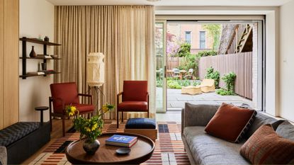 A beautiful living room with a door to the garden and cream curtains. Inside the living room are rust armchairs, gray sofa, black stool, round coffee table, floating shelves, and a floor lamp