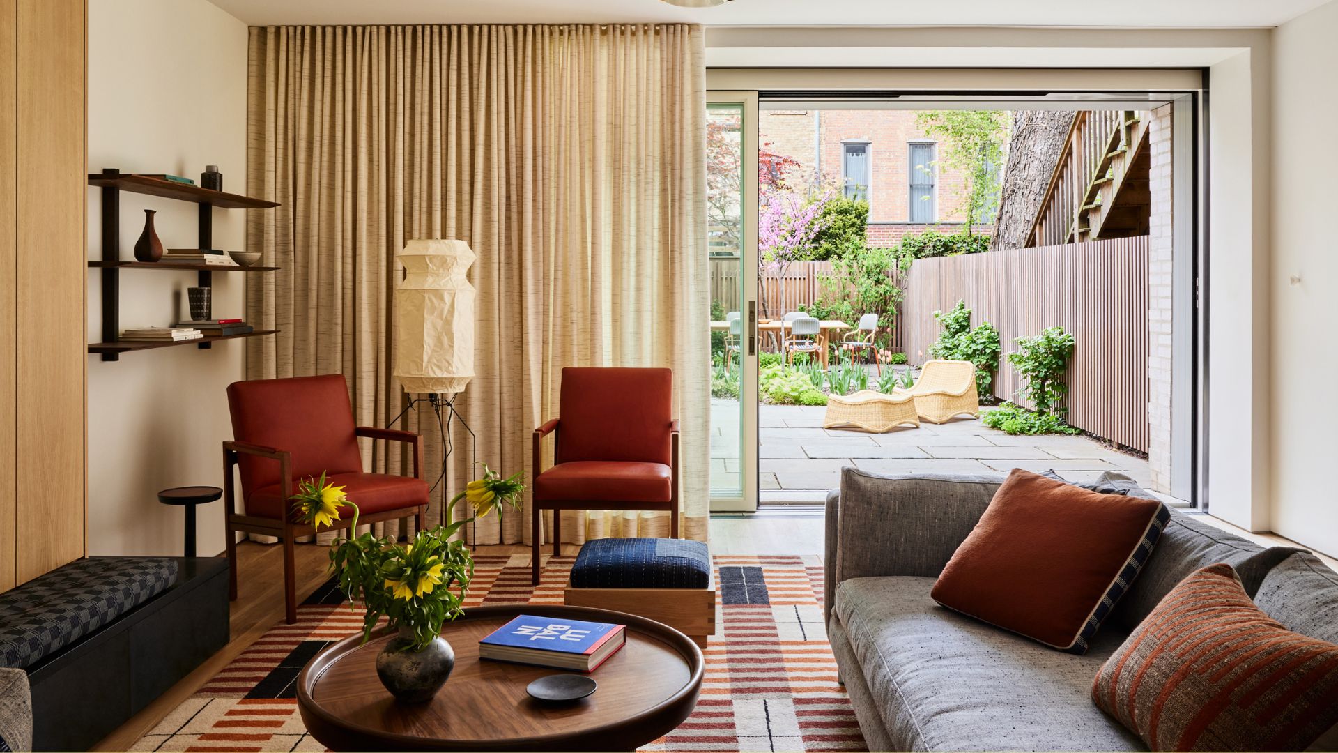 A beautiful living room with a door to the garden and cream curtains. Inside the living room are rust armchairs, gray sofa, black stool, round coffee table, floating shelves, and a floor lamp