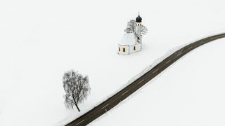 Aerial view of road running through snow-covered field. There is a church and a tree on the left-hand side of the road.