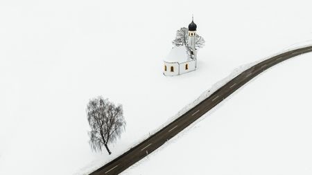 Aerial view of road running through snow-covered field. There is a church and a tree on the left-hand side of the road.