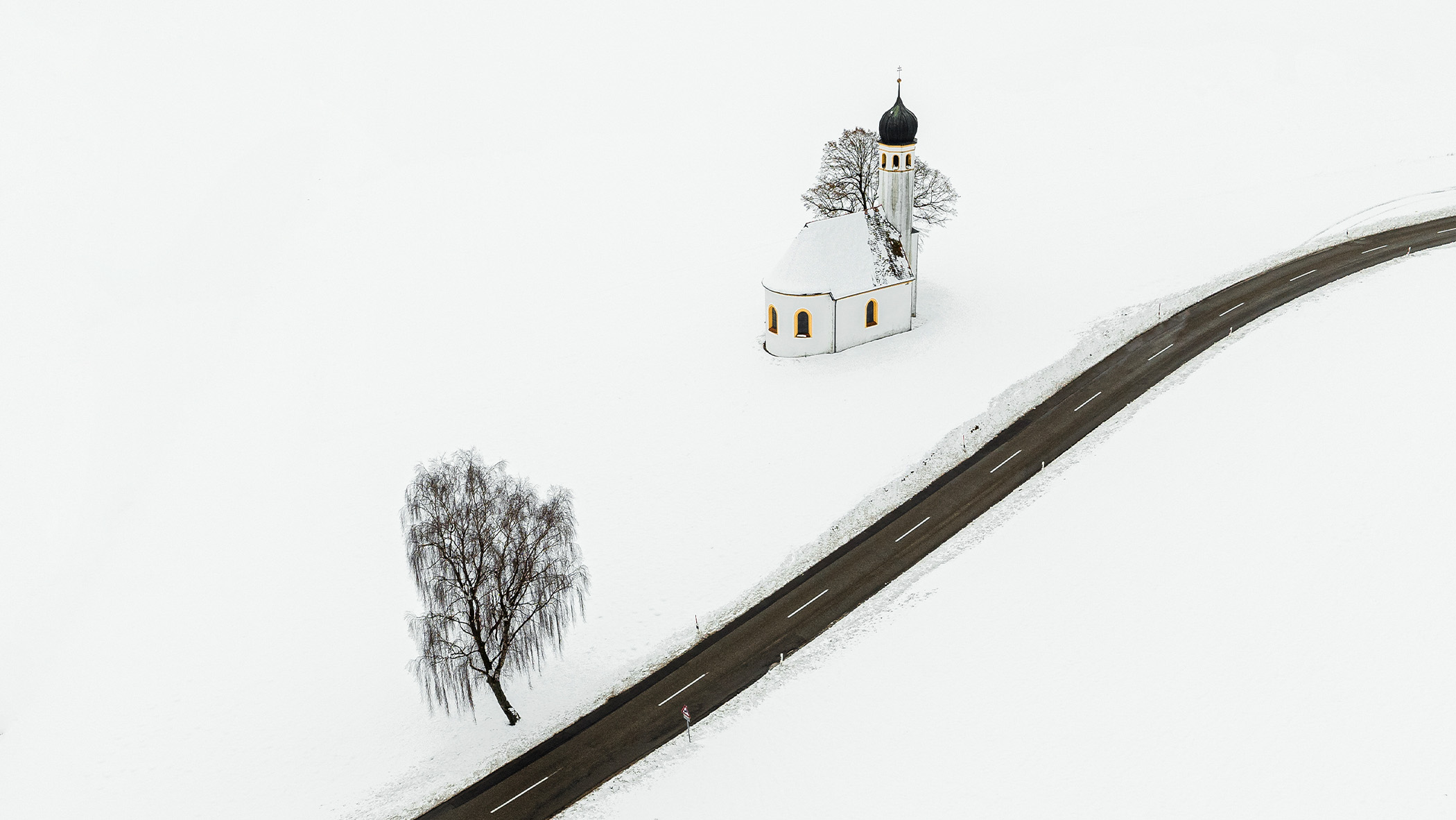 Aerial view of road running through snow-covered field. There is a church and a tree on the left-hand side of the road.