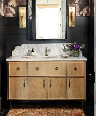 Small black bathroom with large floral wallpaper above vintage vanity, mirror, wall lights, terracotta til