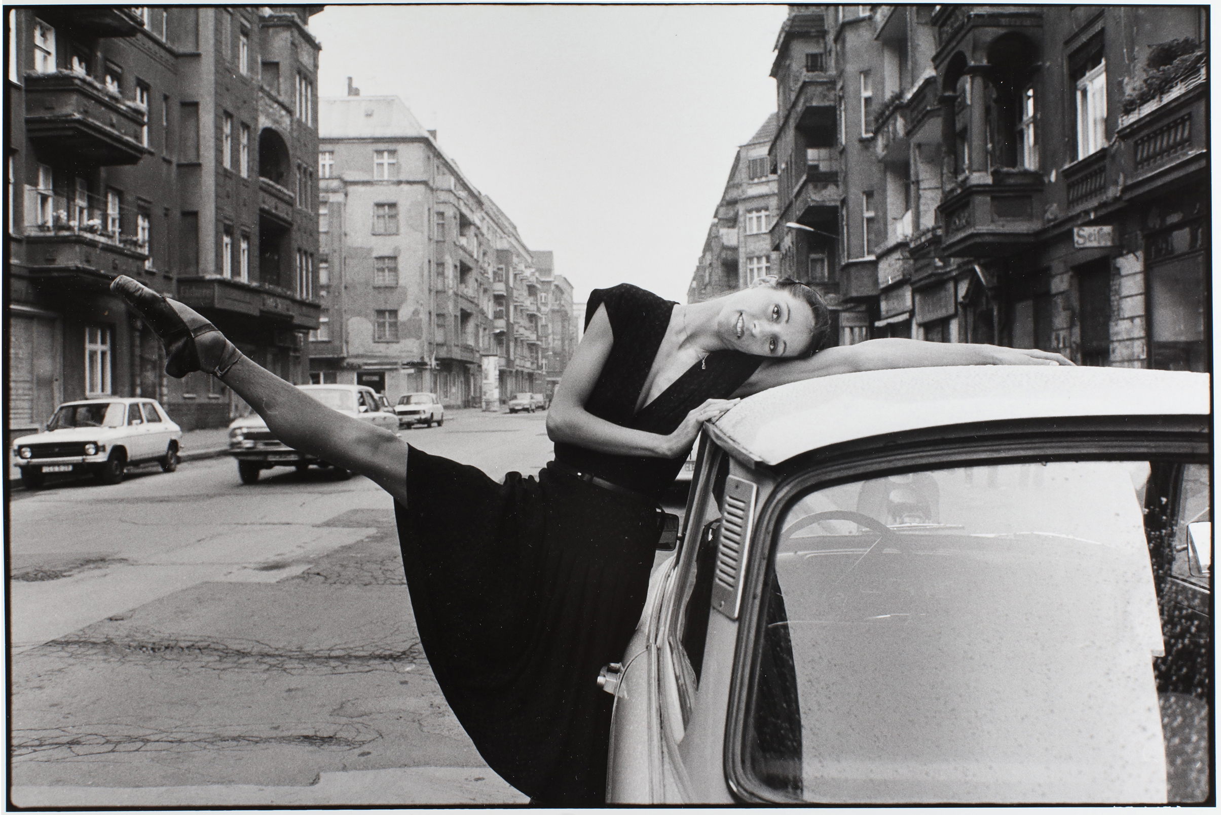 Black and white photo of a woman in a black dress leaning on a car with one leg extended, set on a city street with vintage cars