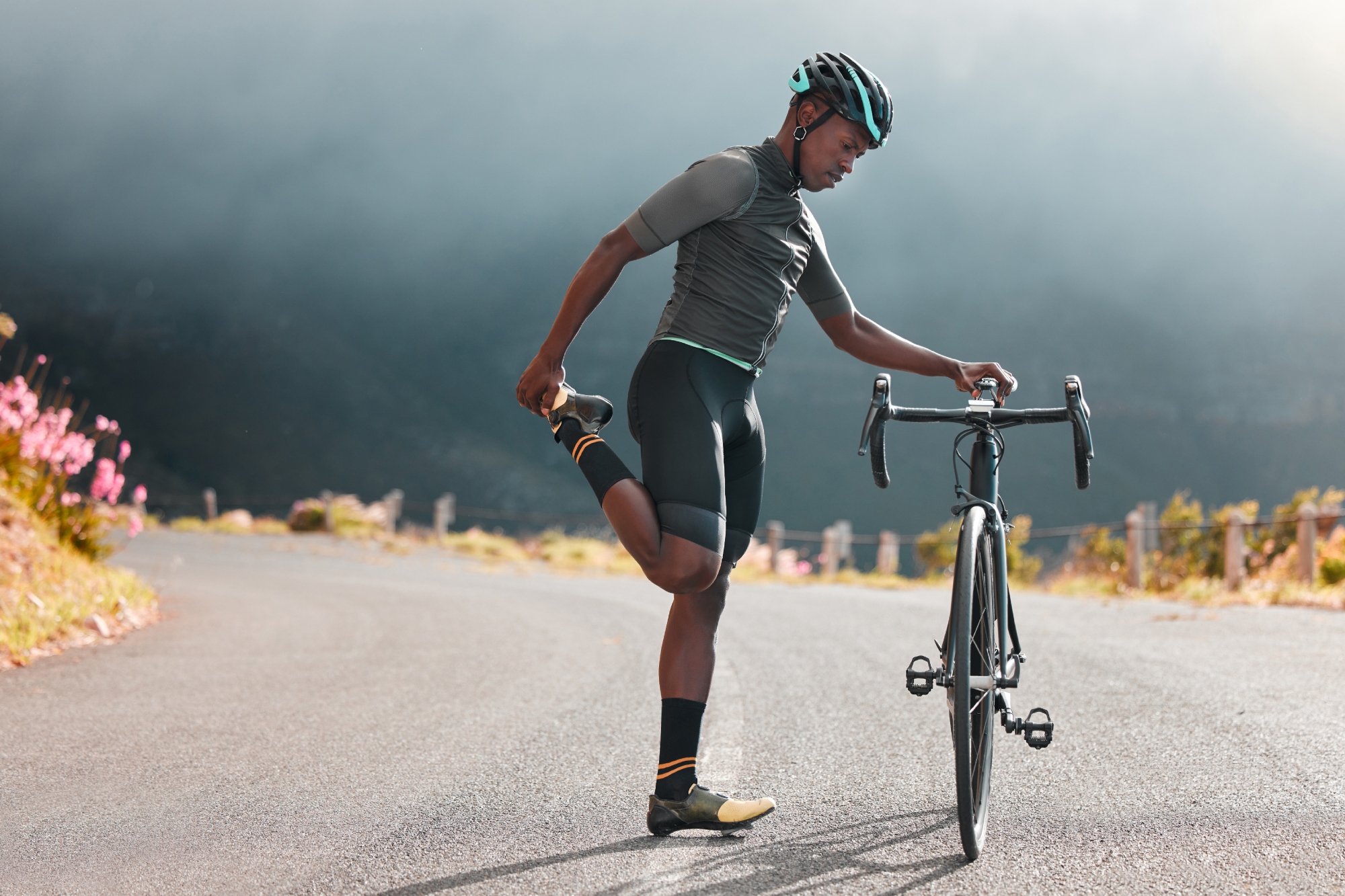 A black bike rider stands streching his quad while holding on to his bike in the road