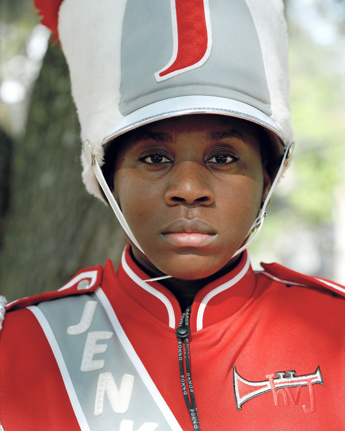 A close-up portrait of a young marching band member in a red jacket and tall white plumed hat, looking straight at the camera