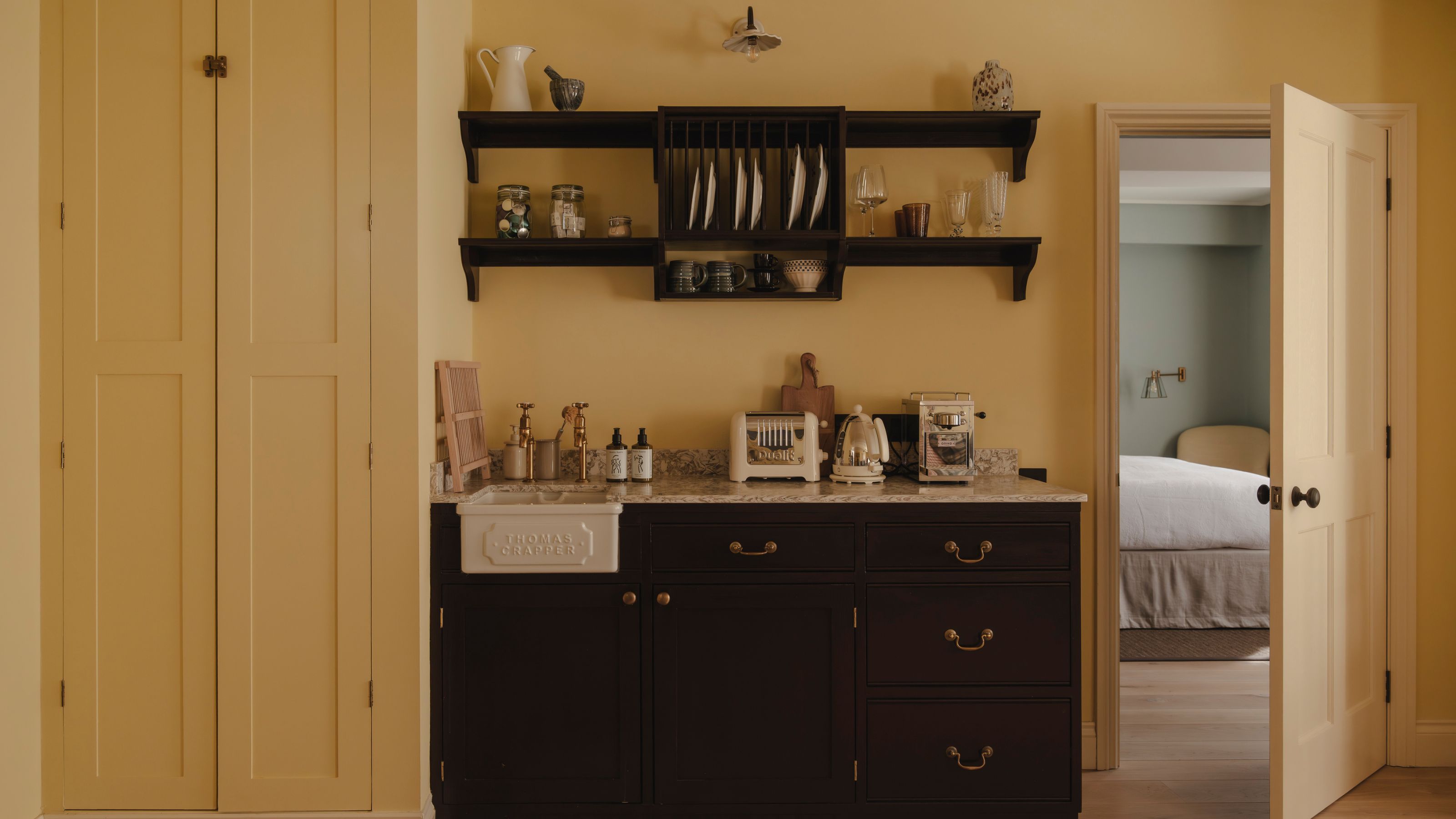 a butter yellow small apartment kitchen with dark kitchen cabinetry, open shelves, a pantry cupboard, and a small thomas Crapper sink