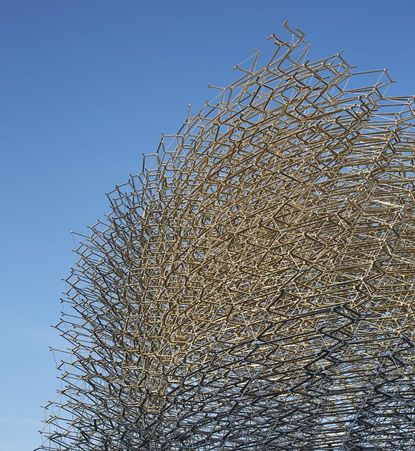 View of the UK Pavilion featuring a tall, aluminium mesh ’beehive’ structure and several patinated metal, geometric planters with low greenery under a clear, blue sky