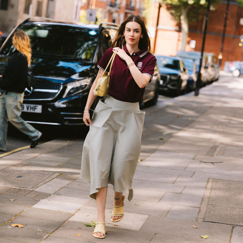 Woman pictured is wearing a gray skirt with an asymmetric hemline, yellow open-toed mules, a burgundy polo style shirt, and a yellow handbag. 