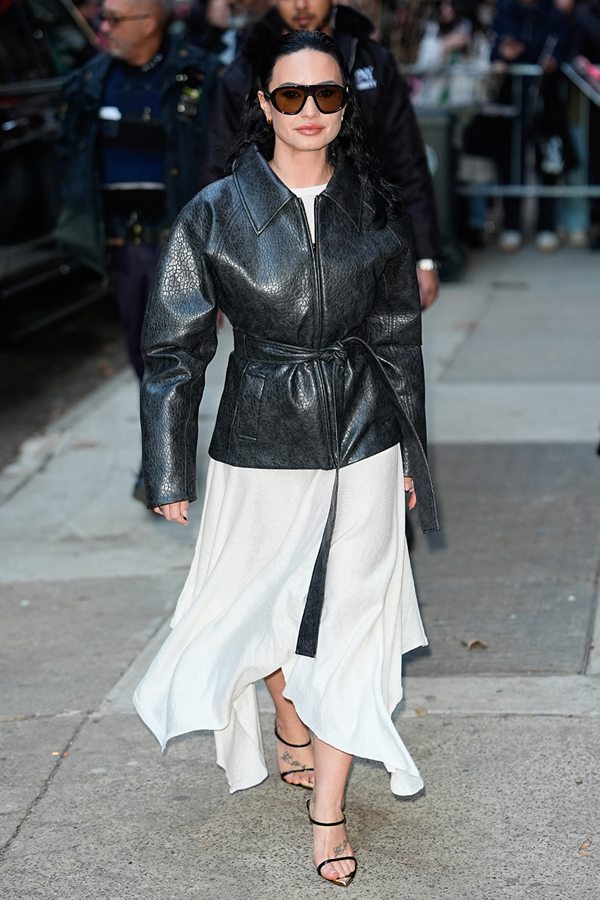 Demi Lovato wearing a belted leather jacket, white skirt, and black sandals