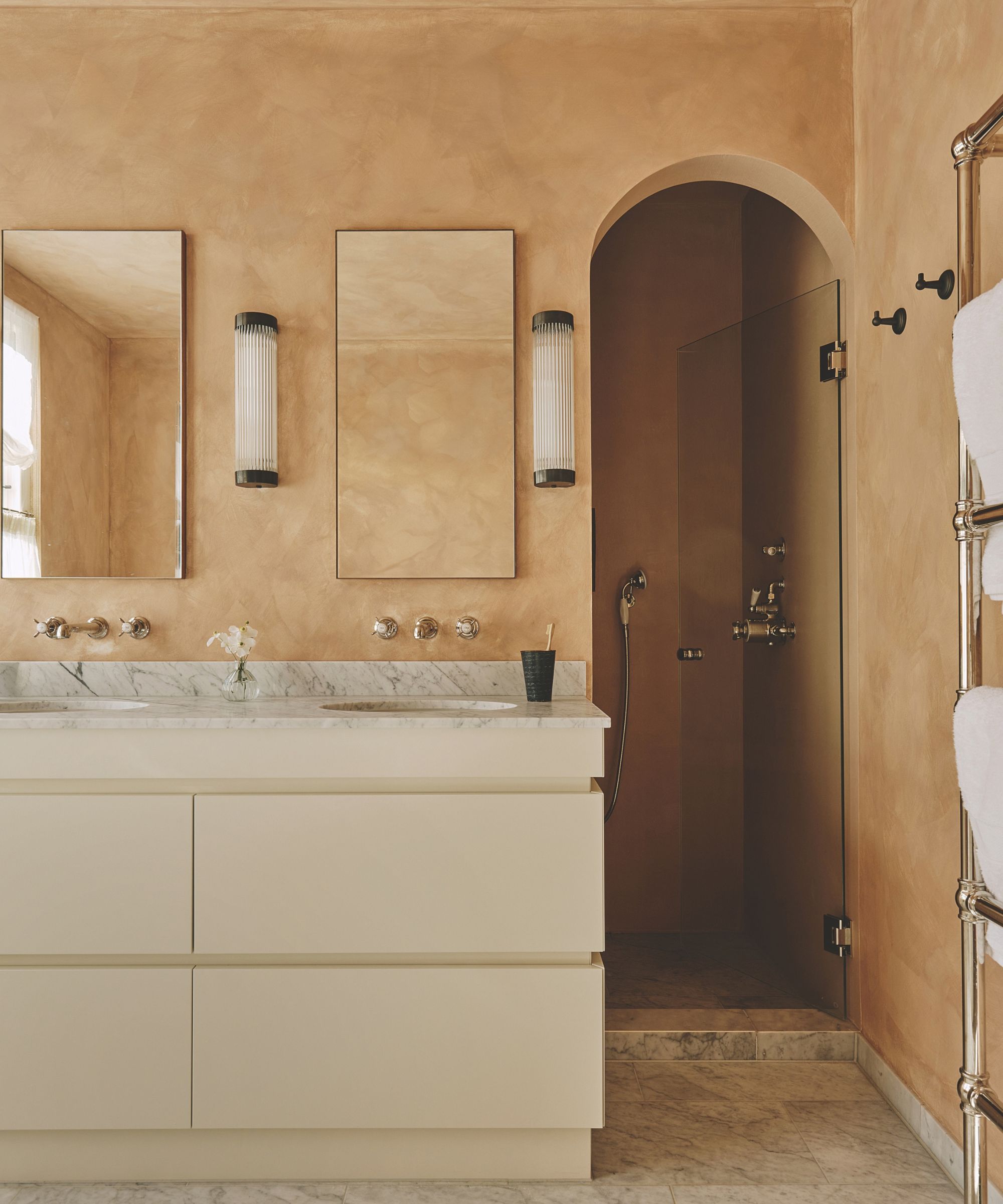 Modern bathroom featuring warm plaster walls, double vanity with marble top, ribbed sconces, wall-mounted mirrors, and an arched shower doorway