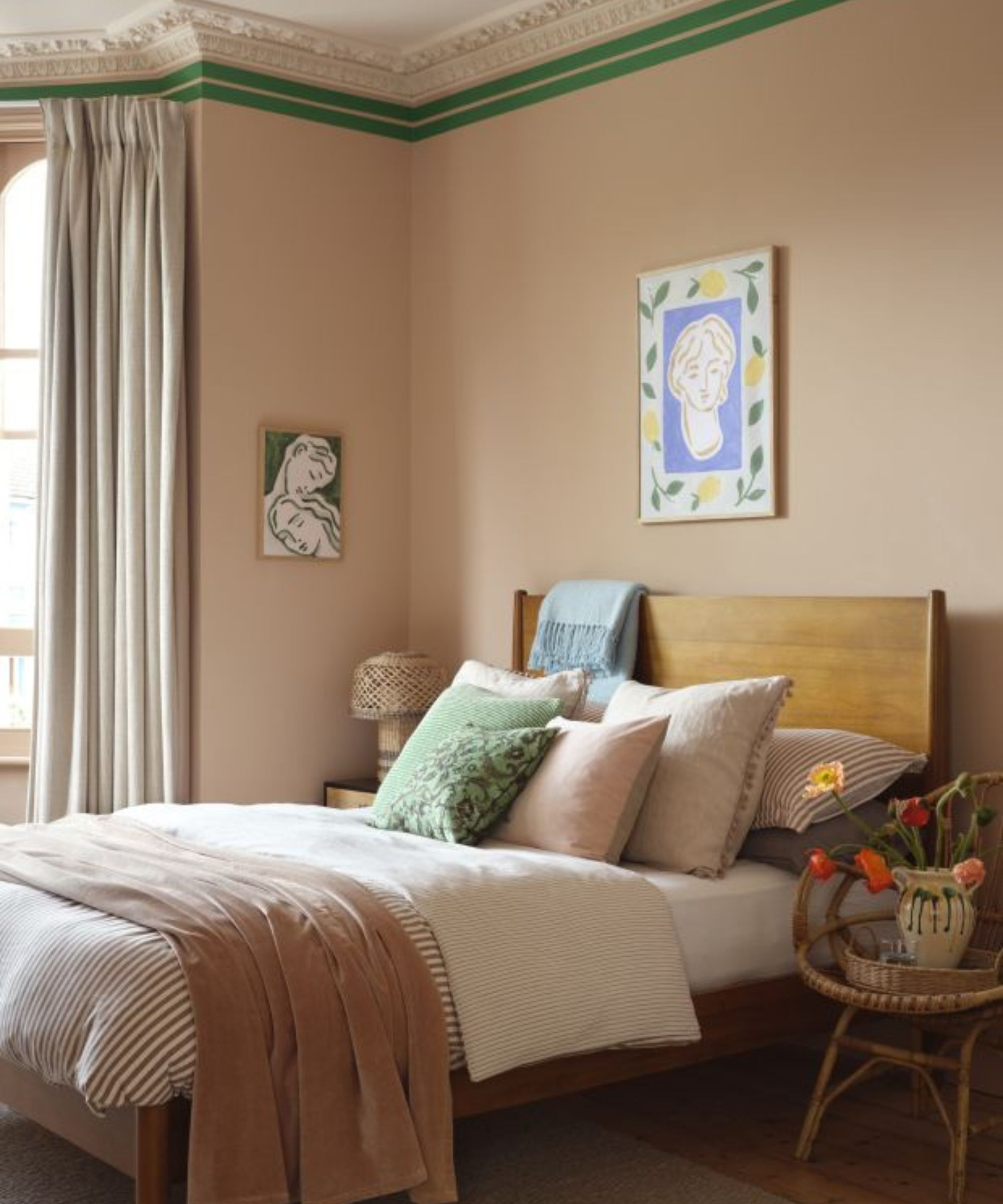Bedroom with high ceilings painted dusty pink and crown moulding painted green, large bed with midcentury wooden headboard, pale pink bedding with velvet throw, and a portrait painting above the bed