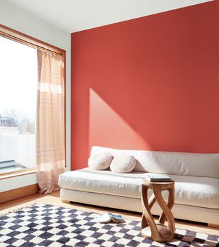 modern living room with one painted soft red/raspberry wall, cream futon style couch, rug, drapes at window, wood side table, balcony