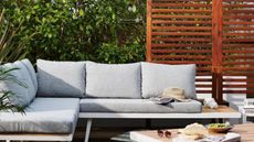 Privacy screen ideas are useful. Here is a backyard decking area with an L-shaped light gay couch with white wooden legs, a light wooden table with bread, olives, and sunglasses on it, and a reddish brown patio screen 