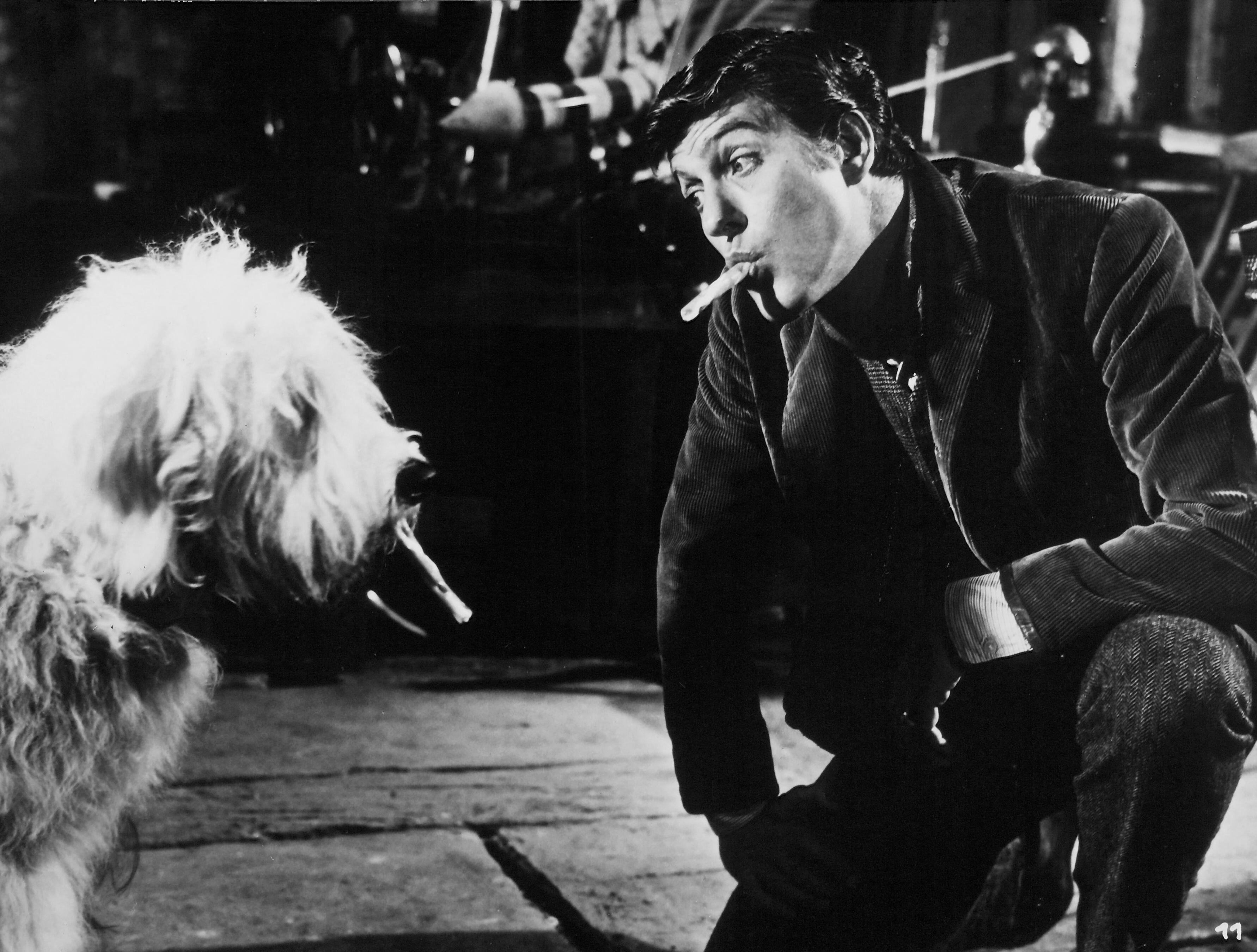 Dick Van Dyke crouches facing a shaggy dog, both holding cigarettes in their mouths in a playful black-and-white behind-the-scenes moment from Chitty Chitty Bang Bang.