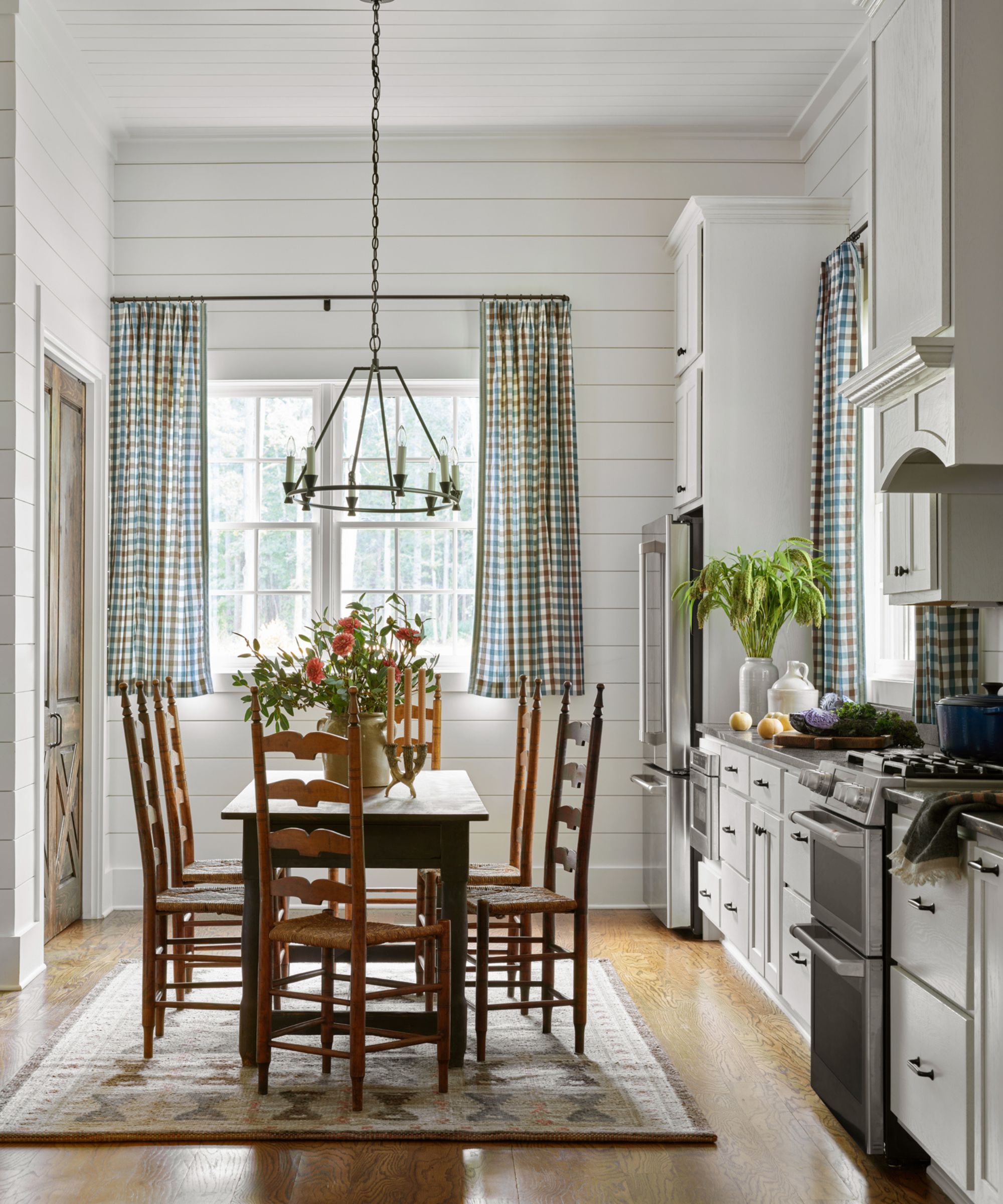 Vintage kitchen with white shiplap walls, vintage wooden dining table and chairs, large rug, gingham curtains and low iron chandelier