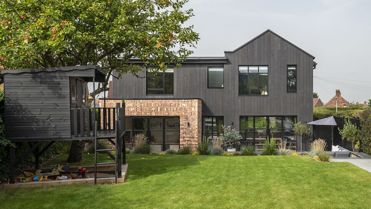 the rear exterior of a black timber clad home renovation and extension project. There is an expansive green lawn in the foreground with a black wooden child&#039;s playhouse to the left