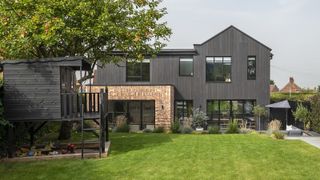 the rear exterior of a black timber clad home renovation and extension project. There is an expansive green lawn in the foreground with a black wooden child's playhouse to the left