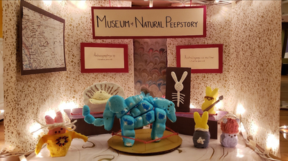 Marshmellow peeps stand around a dinosaur (also made out of peeps) in a scientific diorama. The diorama is titled "Museum of Natural Peepstory"