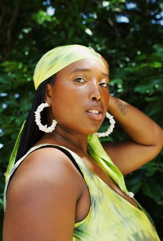 Shell jewelry trend on a woman wearing a green silk scarf over box braids with large shell hoop earrings and a matching green slip dress.