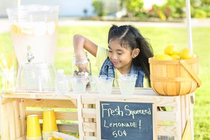 Happy girl makes fresh lemonade to sell at lemonade stand in her front yard. A basket of lemons and a beverage dispenser are on the stand.