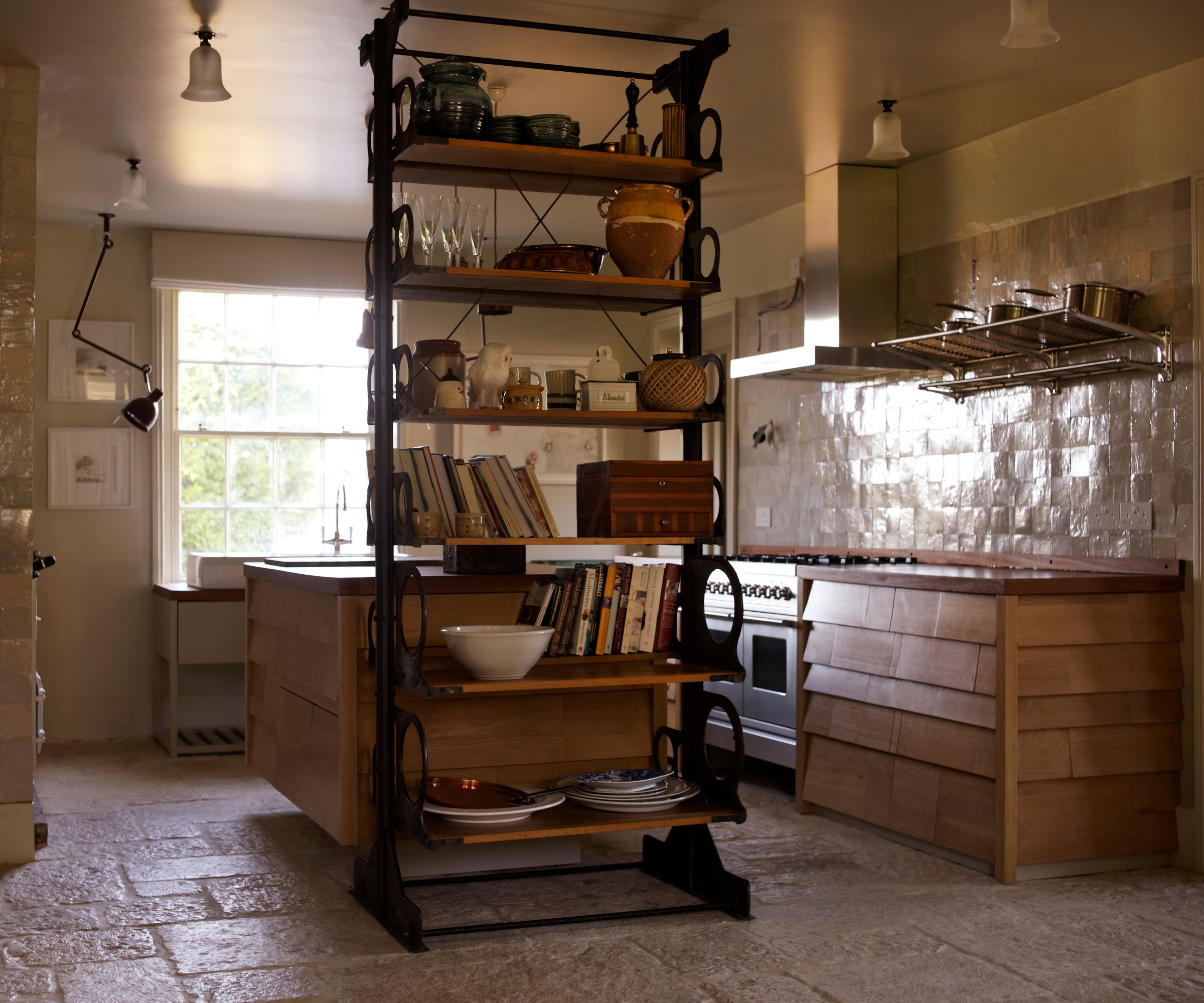 Kitchen with slate floor, cream zeillage tiled backsplash and wooden cabinetry, silver range oven, a freestanding island, and a vintage industrial shelving unit with books, ceramics and tableware.