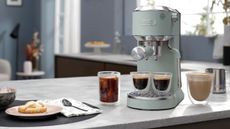 Image of the De'Longhi Dedica Duo machine in sage green on a marble countertop in a blue kitchen. There are two full espresso glasses on the machine and two other coffee drinks beside the machine.