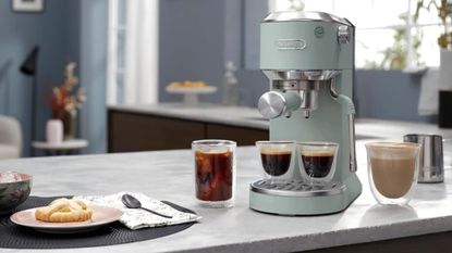 Image of the De'Longhi Dedica Duo machine in sage green on a marble countertop in a blue kitchen. There are two full espresso glasses on the machine and two other coffee drinks beside the machine. 
