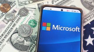 Microsoft Corporation logo seen displayed on a smartphone with United States Dollar notes and coins in the background.