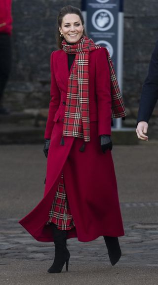 Kate Middleton wearing a long red coat with a red plaid scarf and skirt smiling as she walks down a cobblestone road