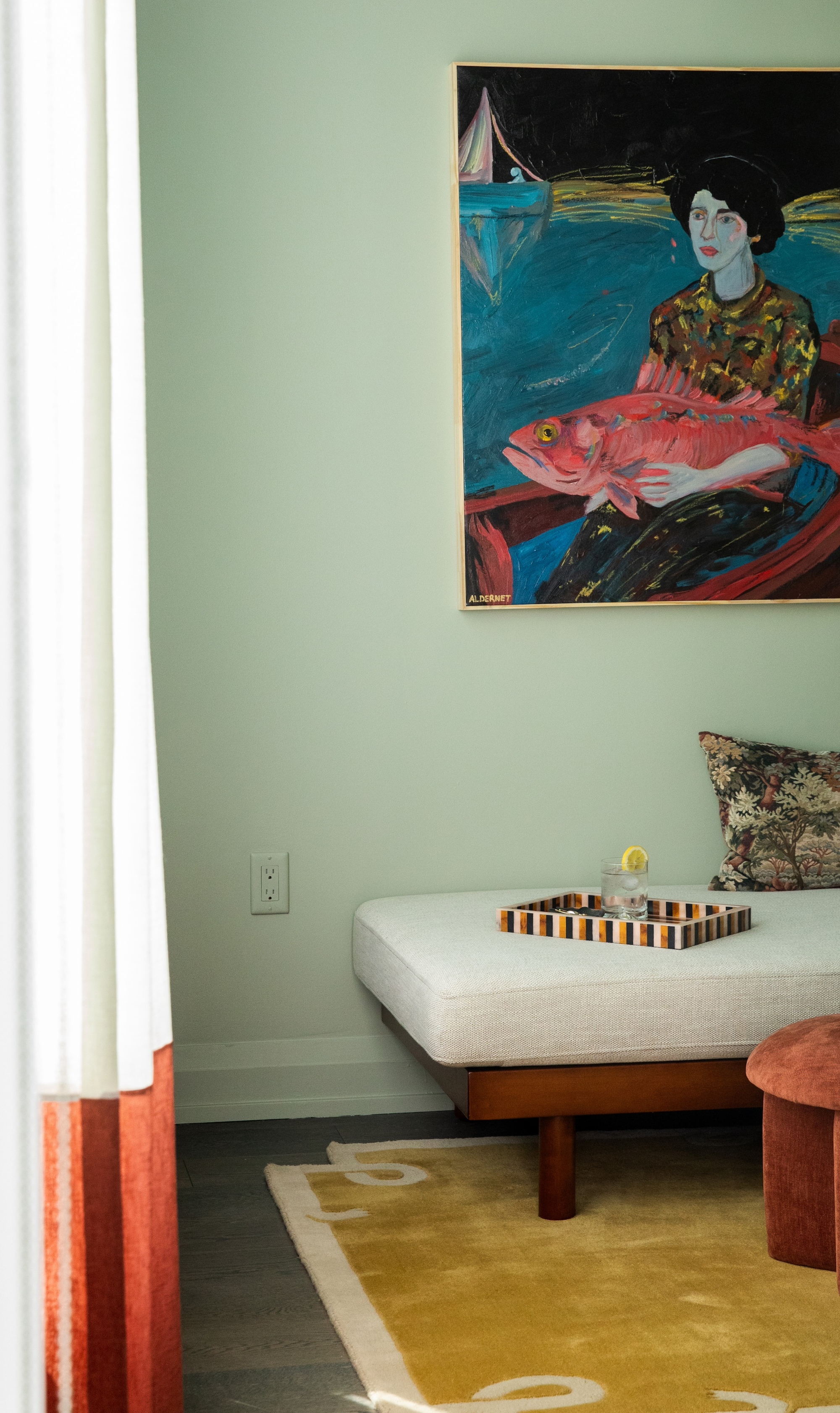 The corner of a sea green living room with a white daybed with a striped tray on it, a yellow rug, and abstract art hanging on the wall.