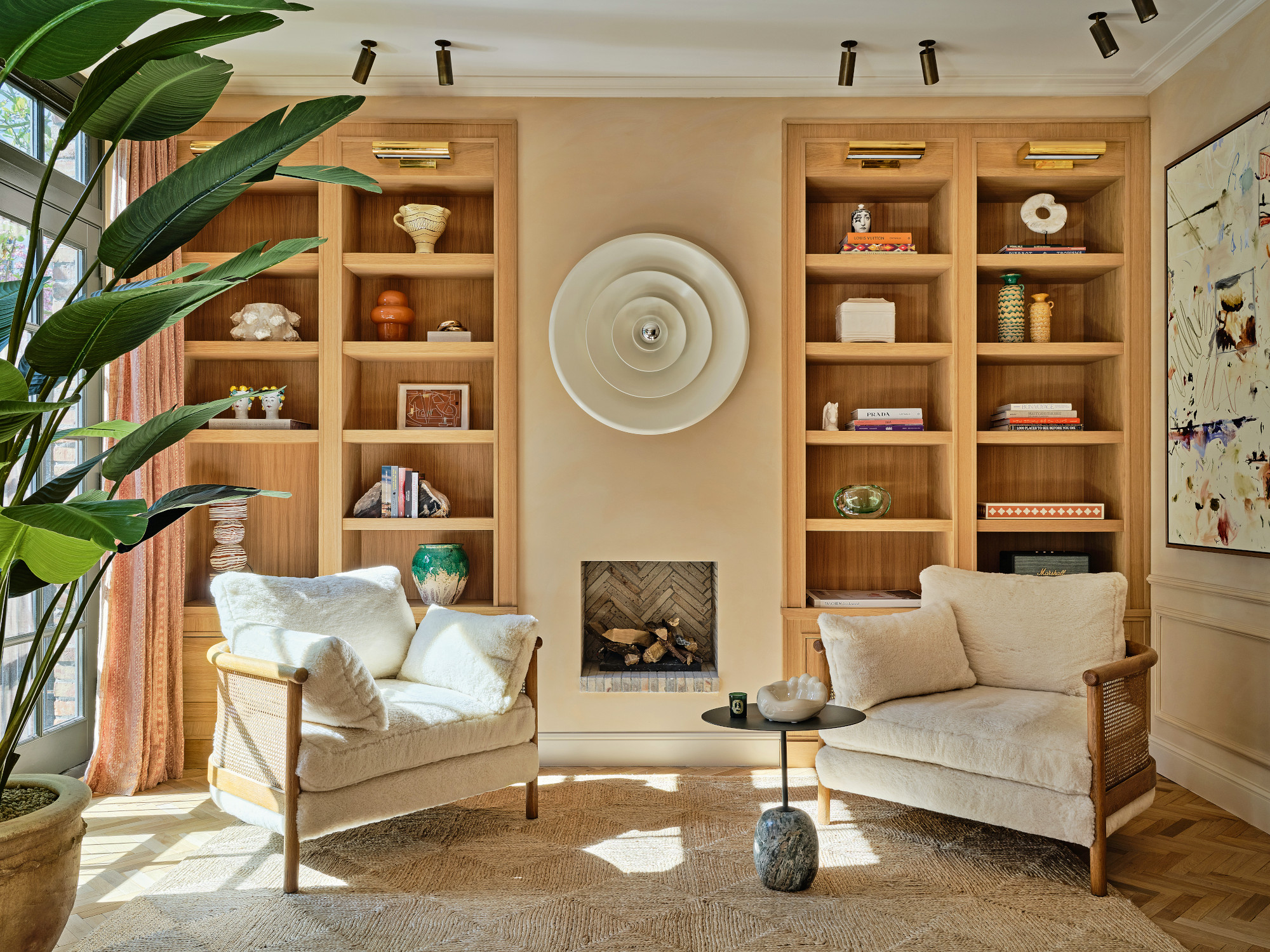 Beige living room with honey oak built-in shelving, cream armchairs with wood and cane frame, jute rug, grey marble side table and white vintage light art piece over inset fireplace