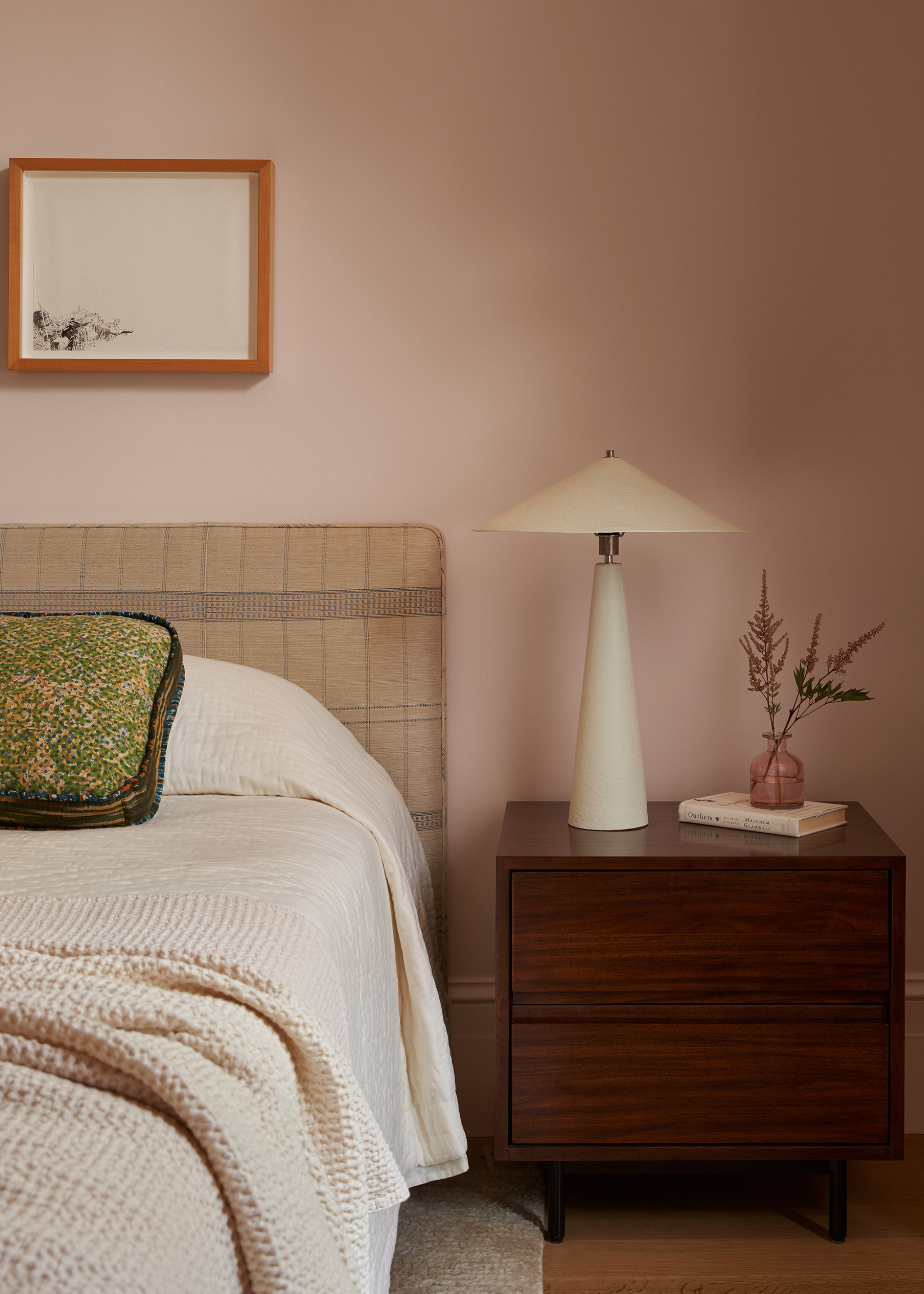 A pale pink bedroom with a wood side table, a white table lamp, a vase of dried stems, a book, all beside a bed with a green pillow, a beige duvet, and a checked headboard