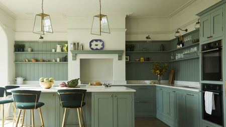 Kitchen island ideas like this one are so chic. Pictured is a sage green kitchen island with marble countertop and vegetables on it, dark green velvet bar chairs with gold legs, two gold pendant lights, and green shelves on white walls behind it