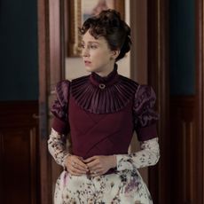 taissa farmiga as gladys russell wearing a maroon and floral dress standing in a parlor in the gilded age