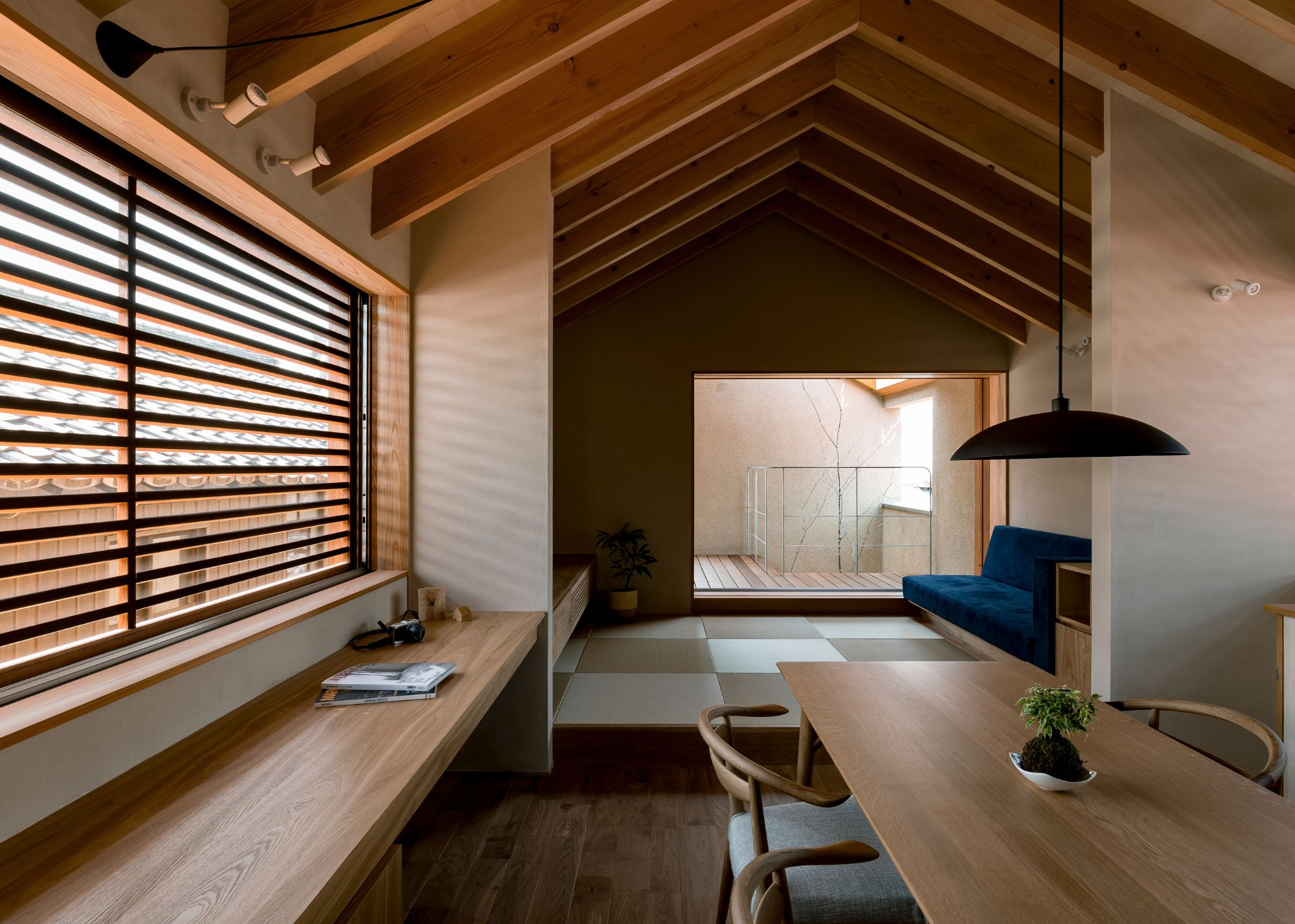 A minimalist Japanese open plan living space with neutral walls and wood frames, with a pitched ceiling, a wood dining table, and a low-profile sofa with a raised large format checkerboard floor