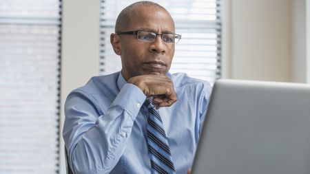 A serious older man wearing a tie looks at his laptop while sitting in his nondescript office. 