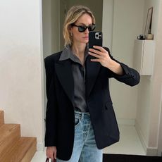 Woman in hallway wears black blazer, grey shirt, blue jeans