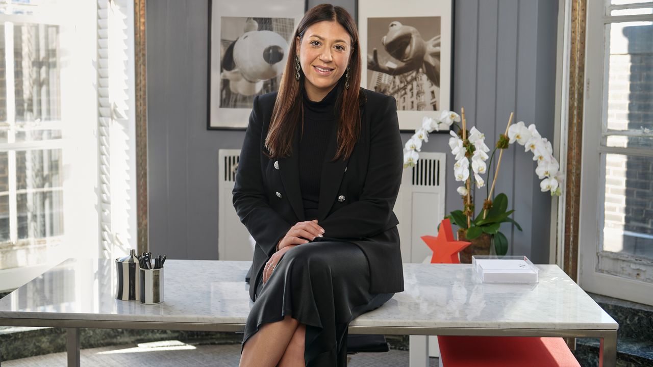 Nata Dvir, Chief Merchandising Officer at Macy’s, in a black dress and blazer sitting on a desk for Marie Claire&#039;s What I Wear to Work franchise.