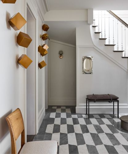Modern hallway with black and white geometric tile, square brass sculptures on wall, gray painted walls and staircase, wooden chair in the foreground