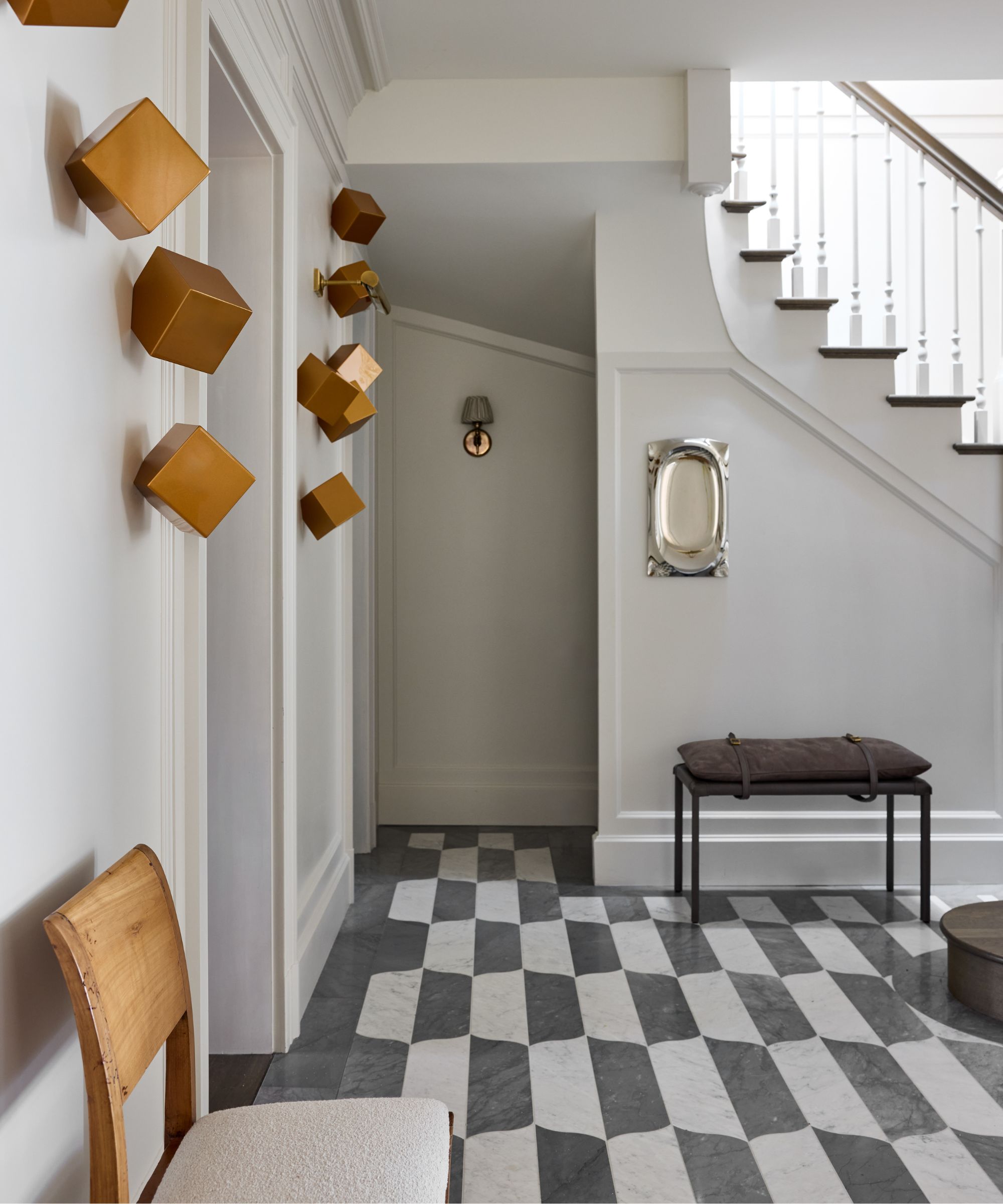 Modern hallway with black and white geometric tile, square brass sculptures on wall, gray painted walls and staircase, wooden chair in the foreground