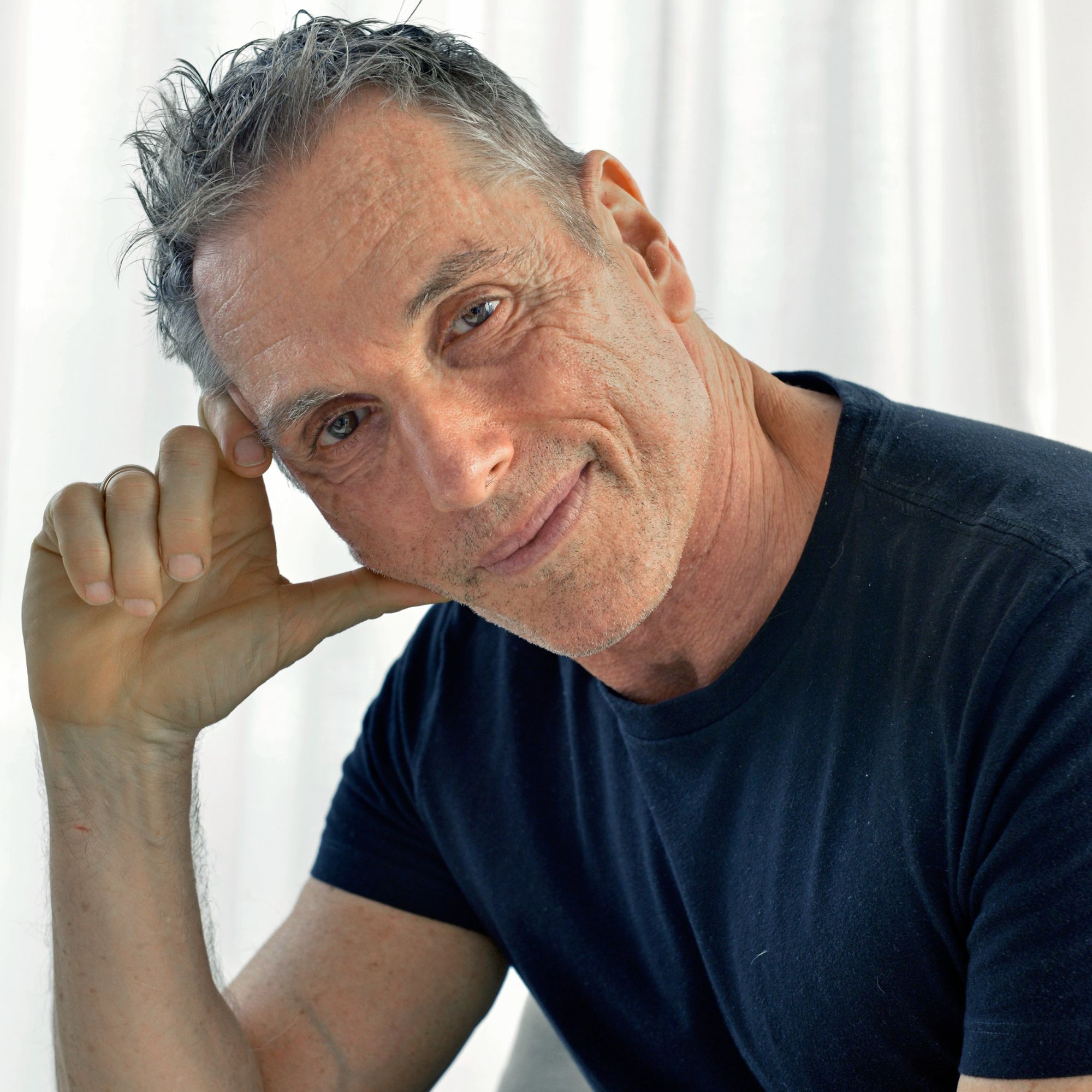 A picture of Vicente Wolf, a man leaning forwards wearing a navy blue t-shirt, with white curtains in the background