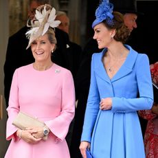 Duchess Sophie wearing a pink dress and feathered tan hat smiling next to Kate Middleton, dressed in a blue long-sleeved coat and matching hat with an updo